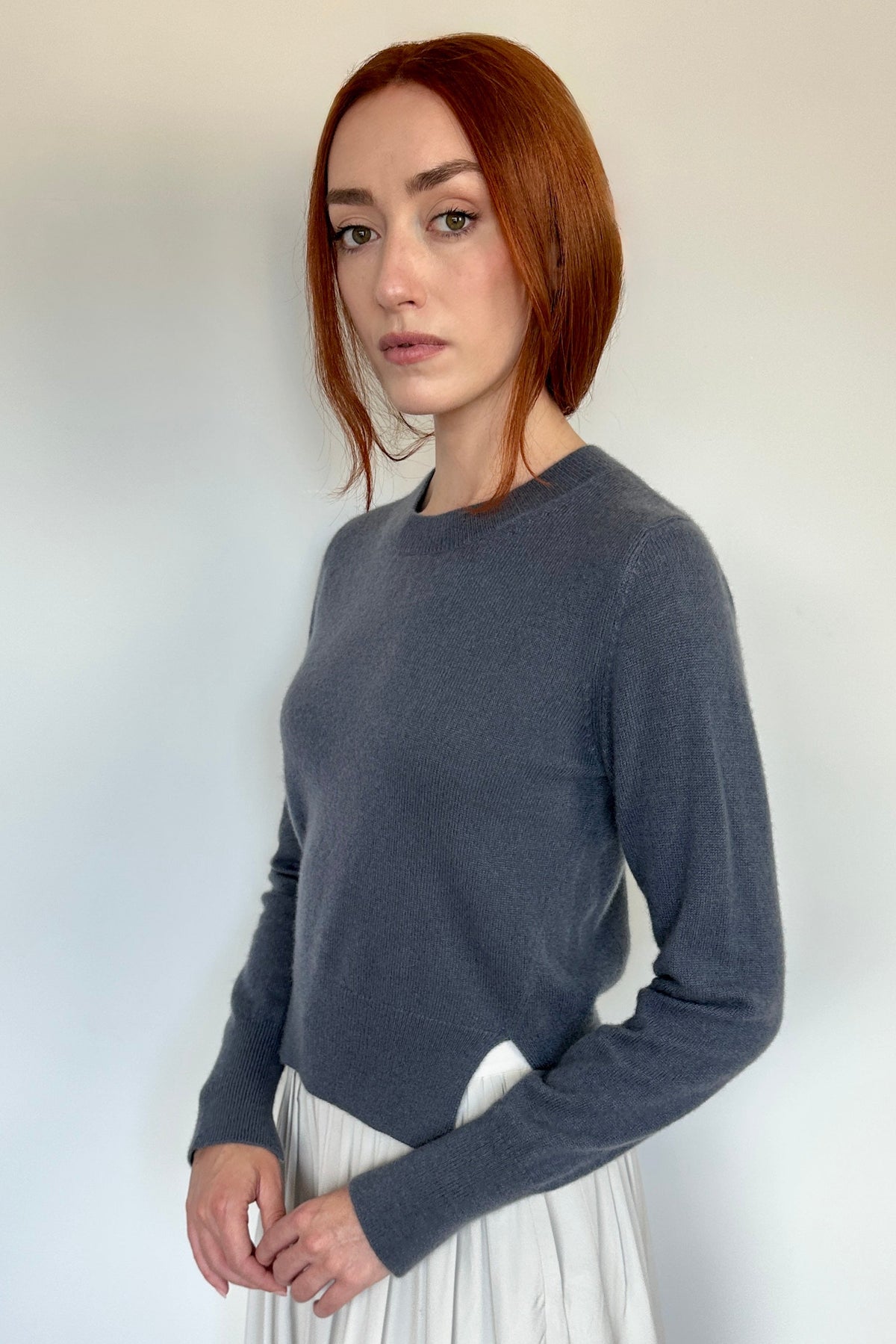 A woman with short red hair stands indoors wearing a dark charcoal gray long-sleeve knit top and a light beige high-low skirt.