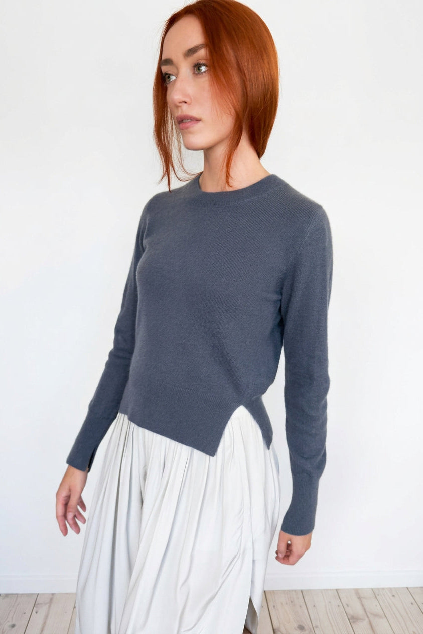 A woman with red hair wearing a dark slate gray long-sleeve knit sweater and a pleated white skirt, standing against a plain off-white wall.