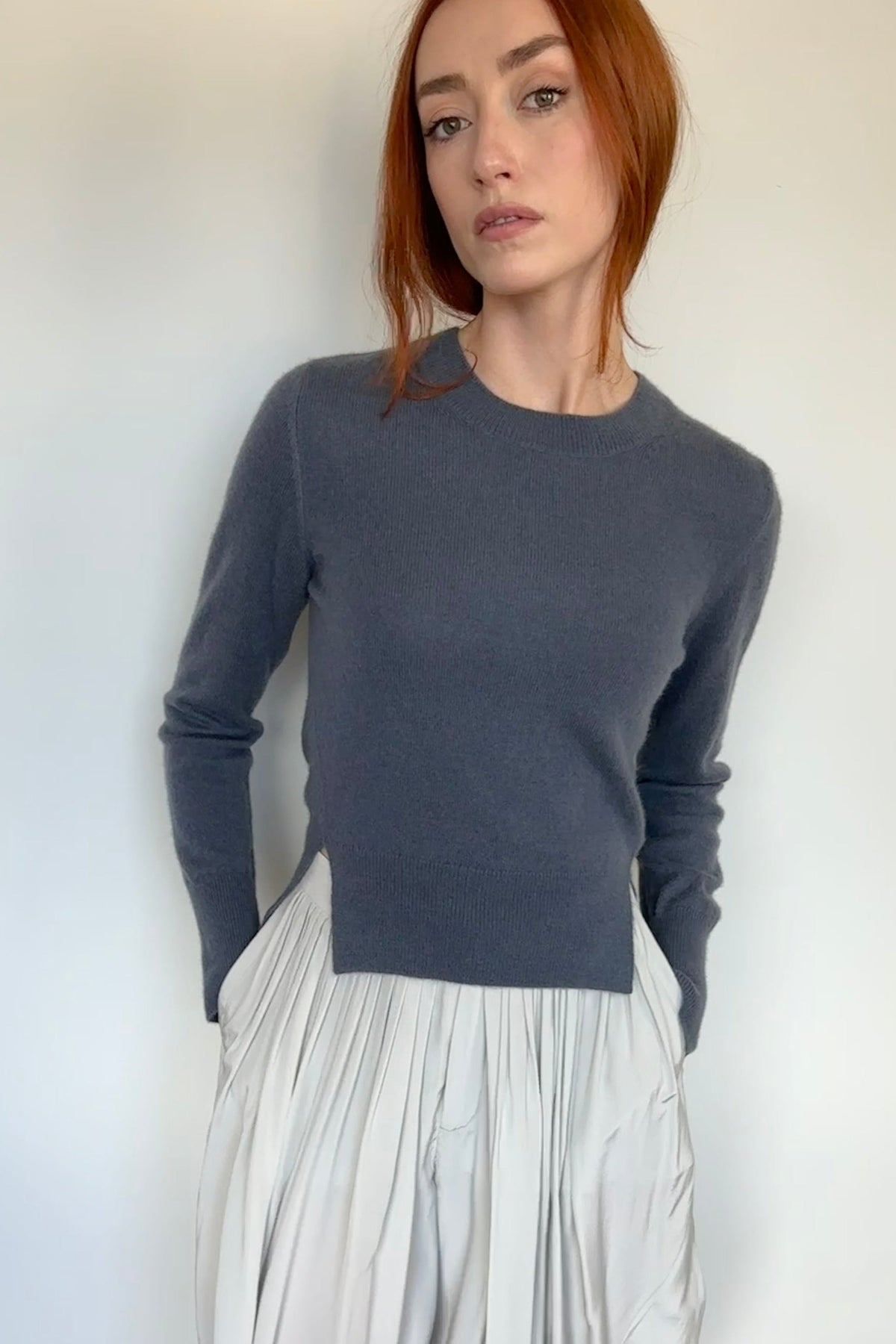 A woman with red hair wearing a dark gray long-sleeve knit sweater and a light gray pleated skirt, standing against a white wall on a light wood floor.