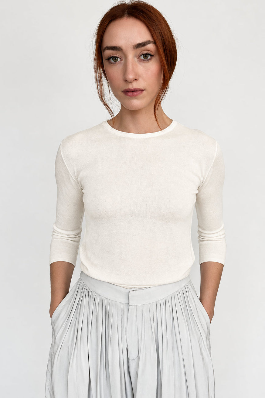 A woman modeling a cream-colored, three-quarter sleeve crewneck top paired with light gray pleated trousers, set against a plain light gray studio background.