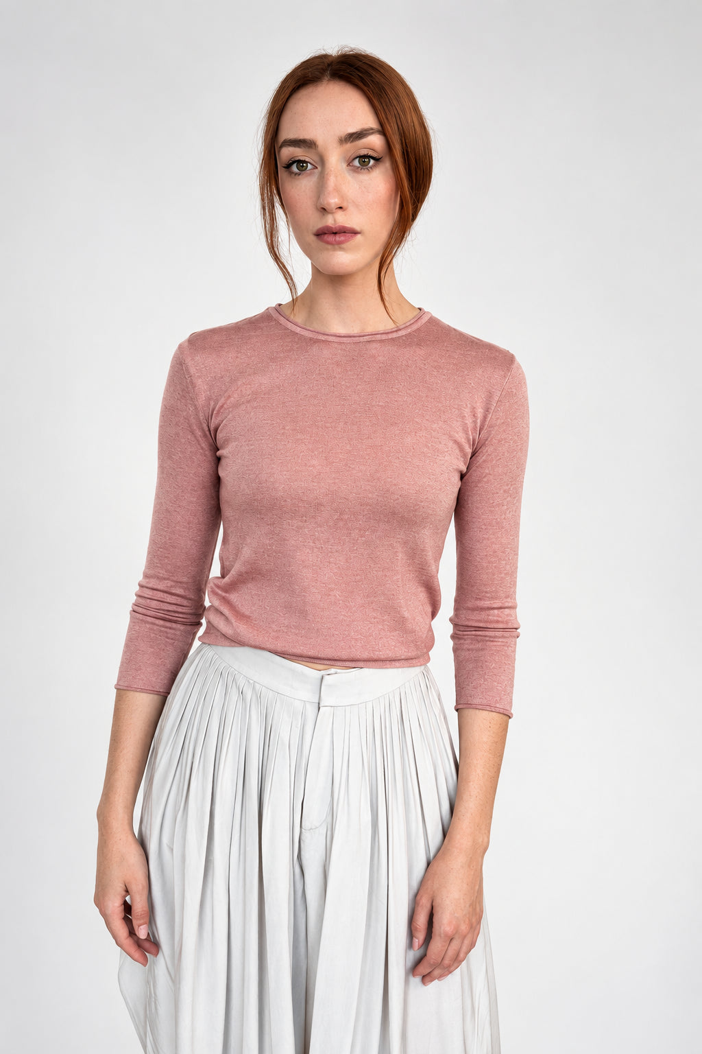 A woman with red hair stands against a plain light gray wall wearing a dusty rose ribbed knit long-sleeve top and white pleated trousers.