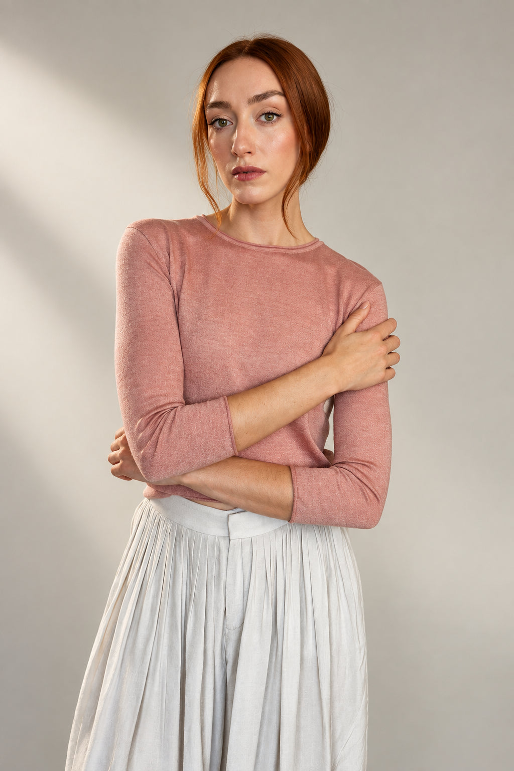 A woman with red hair wearing a dusty rose pink long-sleeve knit top and a light gray pleated skirt, standing against a plain off-white wall.