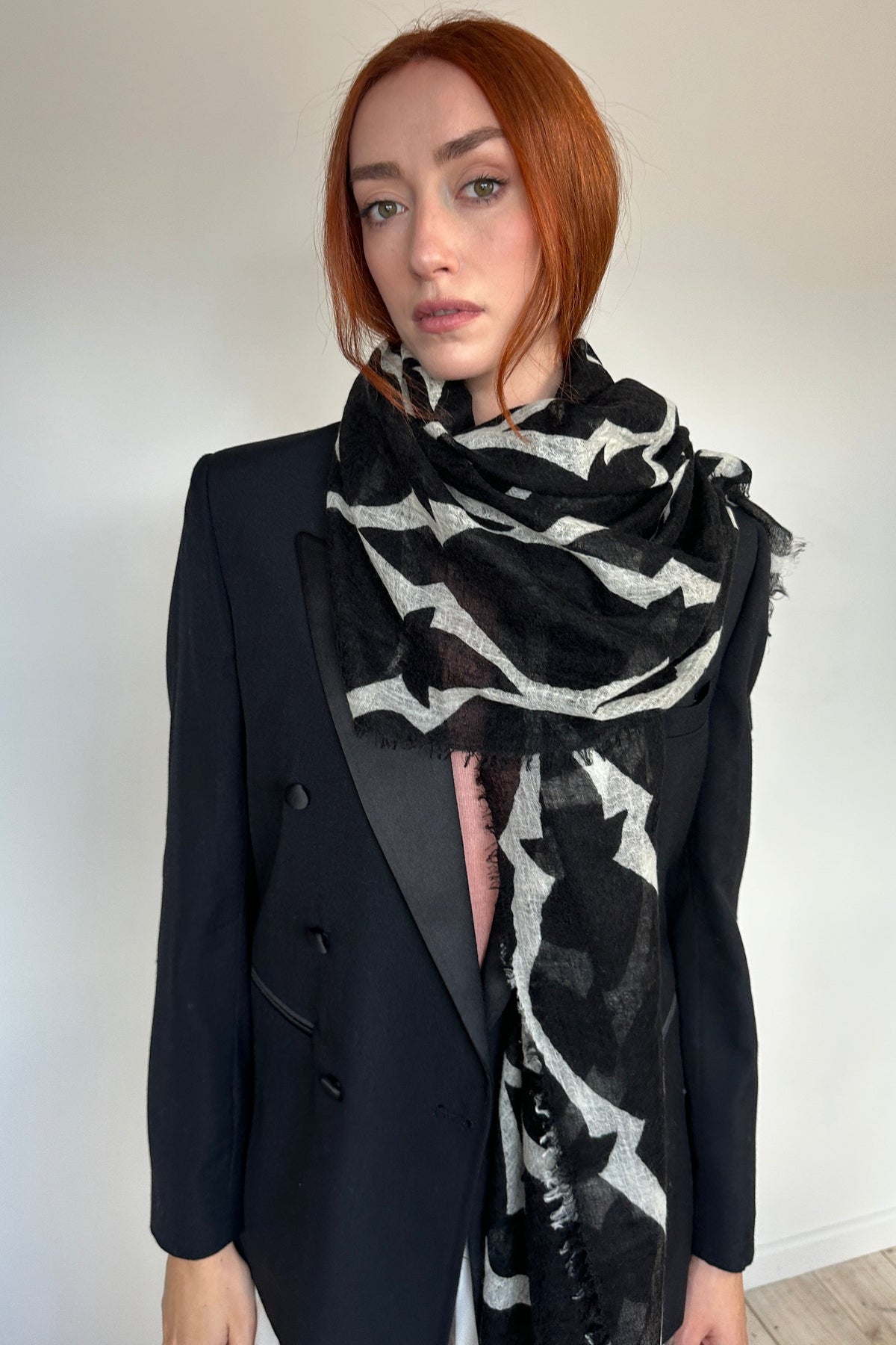 A model with red hair wearing a black double-breasted blazer and a black and white patterned scarf, standing against a plain off-white wall.