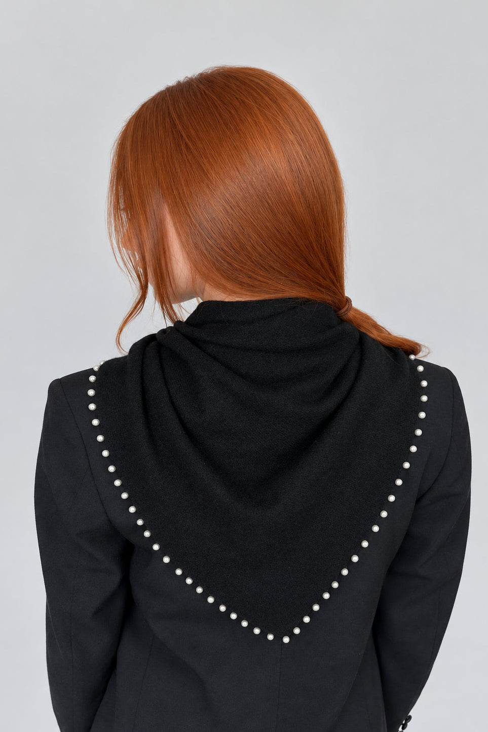 A person with red hair seen from behind, wearing a black hooded jacket with white pearl-like stud embellishments along the hood's edge, against a plain light background.