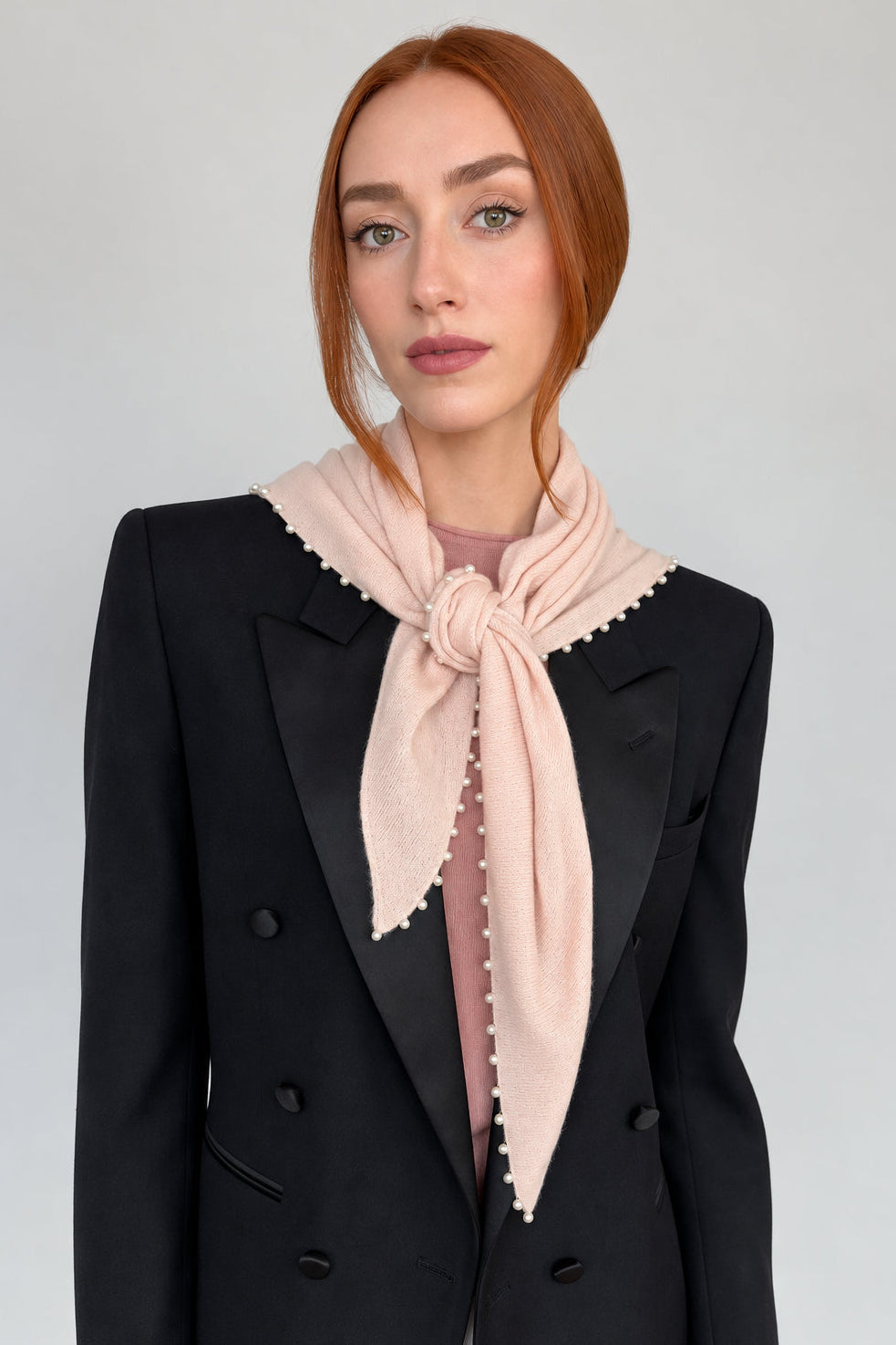 A woman with red hair wearing a black double-breasted blazer with satin lapels, a dusty pink top, and a light pink scarf with white pearl edges tied around her neck, set against a plain light gray background.