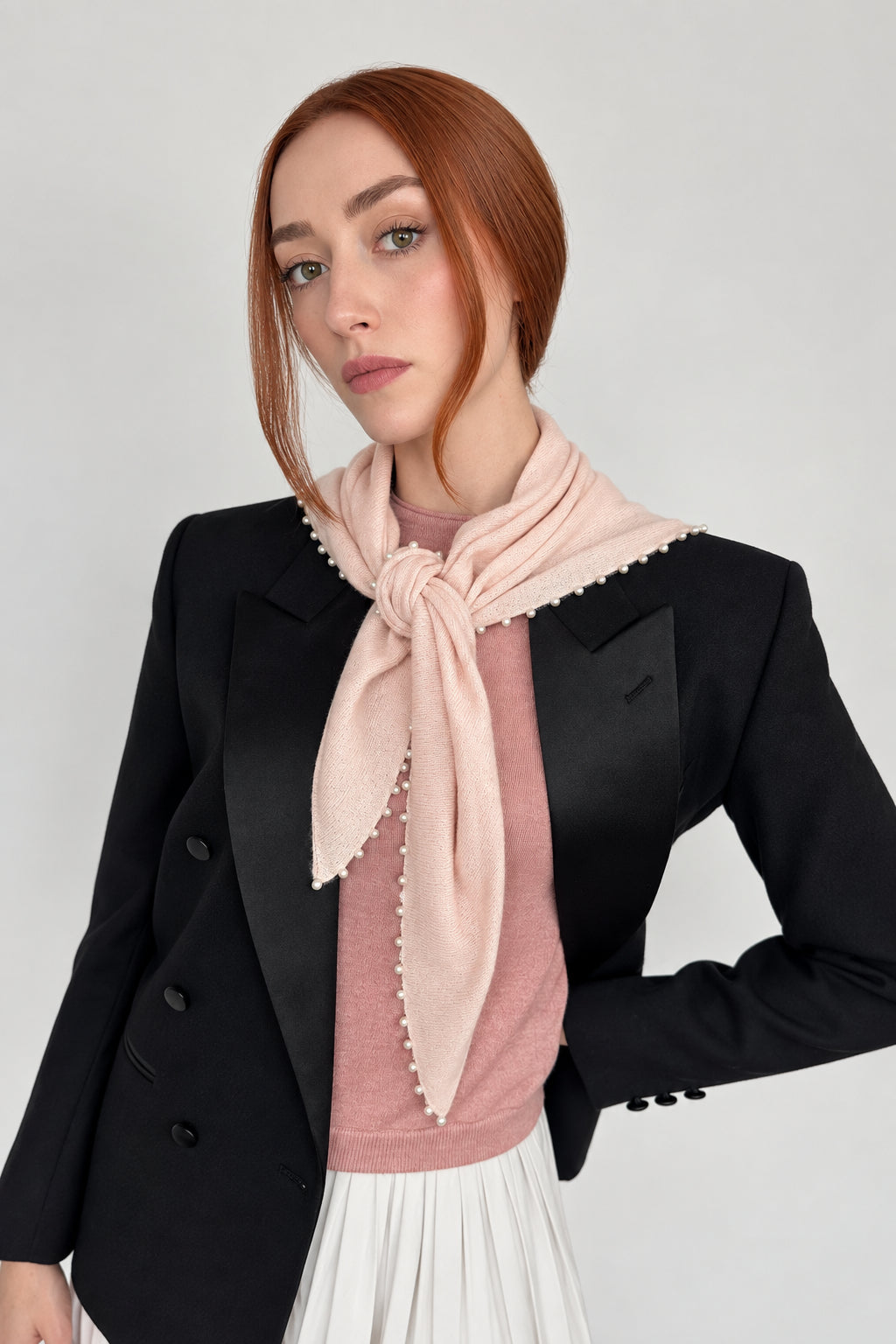A woman with red hair wearing a black blazer, a dusty pink knit top, and a light pink scarf with white pearl edges, posed against a plain off-white wall.
