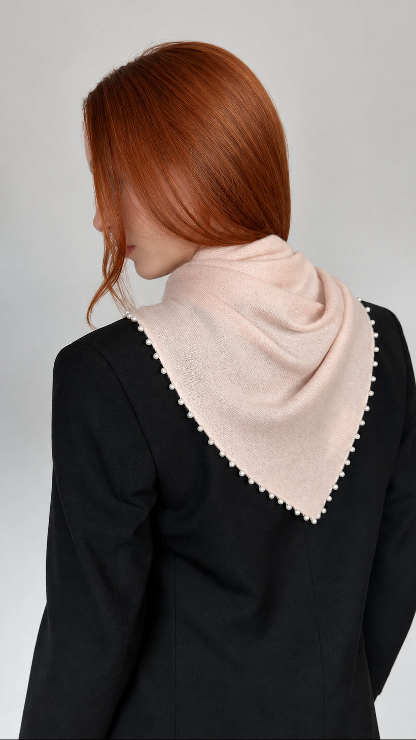 A woman with red hair is shown from behind wearing a light pink knit cowl scarf with a white pearl trim, styled over a dark navy blazer against a plain light gray background.
