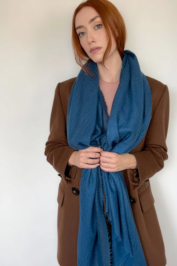 A woman with red hair wearing a brown blazer, white skirt, and a blue textured scarf, standing against a plain off-white wall.
