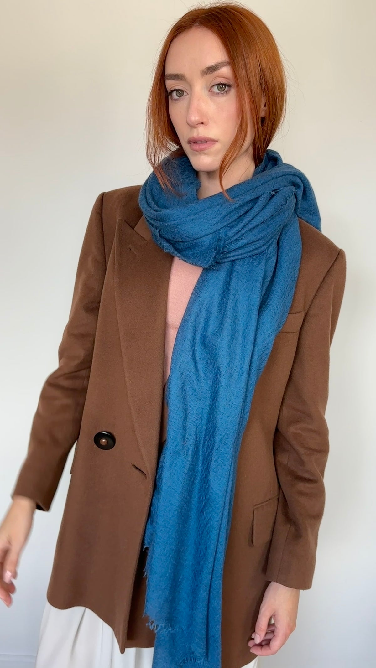 A woman with red hair wearing a brown blazer, a light pink top, and a bright teal blue knit scarf, standing against a plain off-white wall.