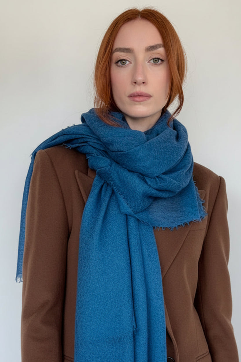 A woman with red hair wearing a brown coat and a bright peacock blue fringed scarf, standing against a plain light gray background