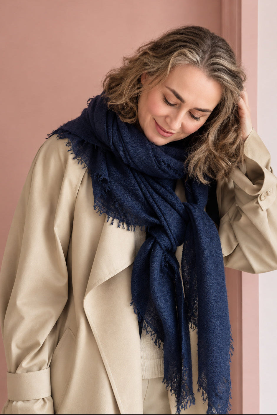 A woman wearing a beige trench coat and a dark navy blue scarf, standing in front of a dusty pink door.