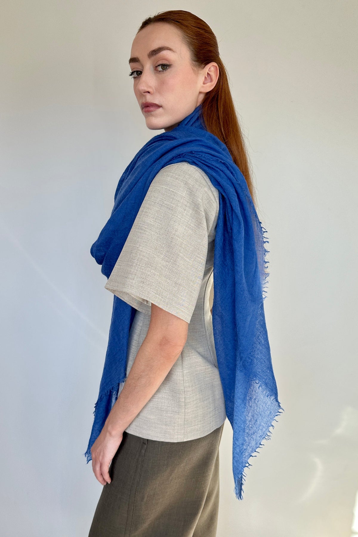 A woman with red hair tied back is shown from the side, wearing a vibrant blue cashmere fringed scarf wrapped around her shoulders over a light gray linen shirt and brown trousers.