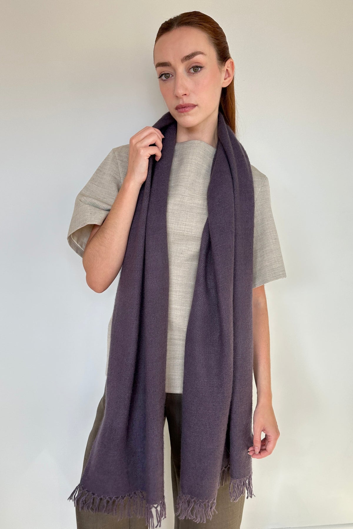 A woman modeling a long, dark purple cashmere fringed scarf draped over her shoulders, paired with a light gray t-shirt and olive green pants, standing against a plain white wall.