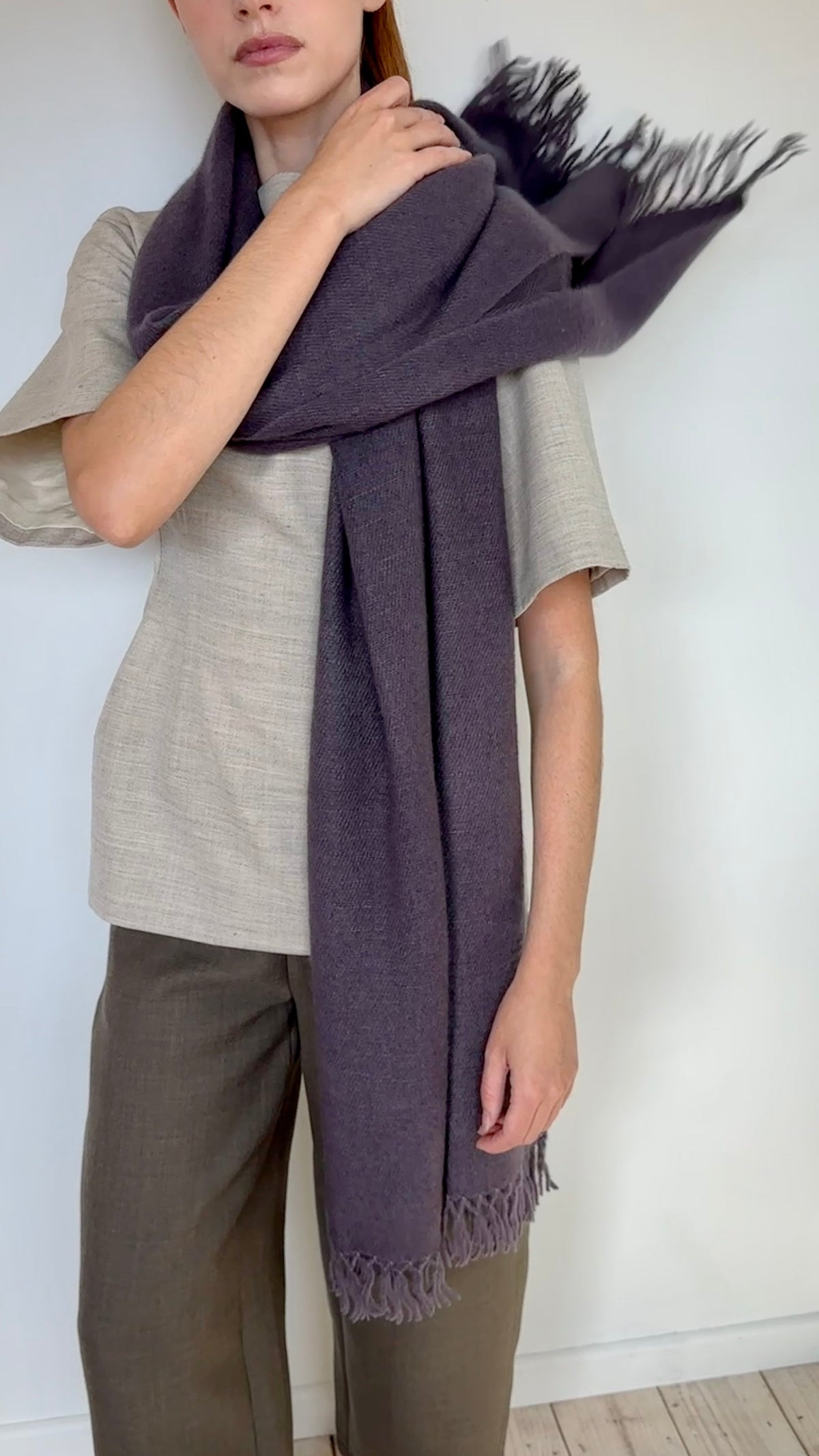 A person wearing a taupe short-sleeve linen top and olive green trousers, wrapped in a dark plum purple cashmere fringed scarf.
