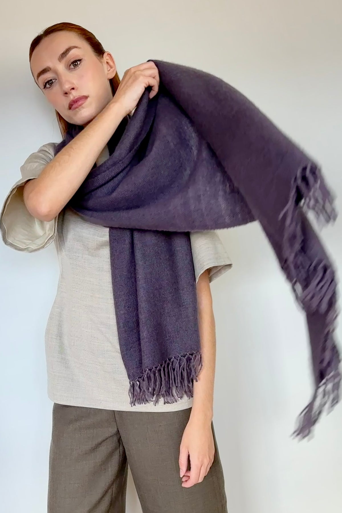 A woman modeling a large, soft woven scarf in a muted plum purple cashmere color, draped over her shoulders and held out to show its fringed edges against a plain light background.