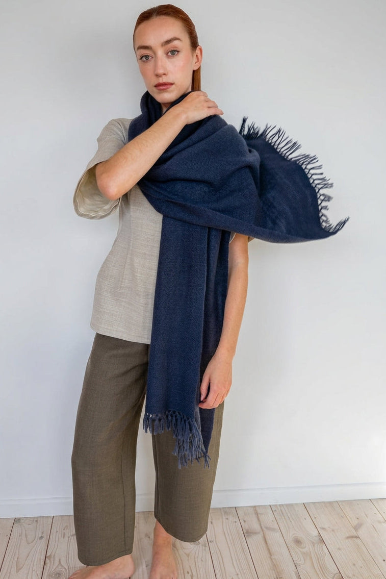 A woman modeling a dark navy blue fringed scarf wrapped around her shoulders, paired with a beige top and olive green pants, standing against a plain off-white wall on a light wood floor.
