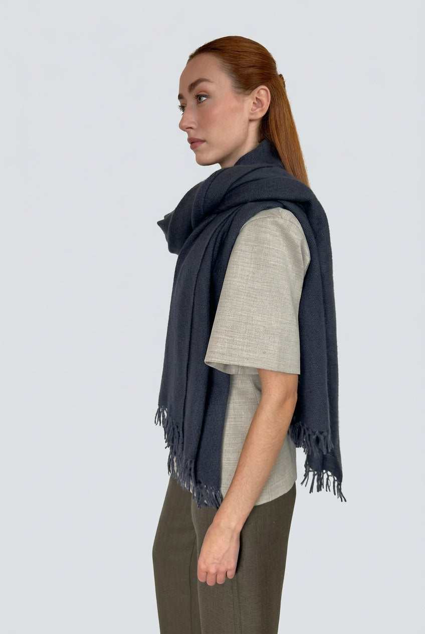 A woman with red hair tied back is shown in profile wearing a dark navy blue fringed scarf wrapped around her shoulders over a light beige short-sleeve top and brown pants, standing against a plain light gray wall.