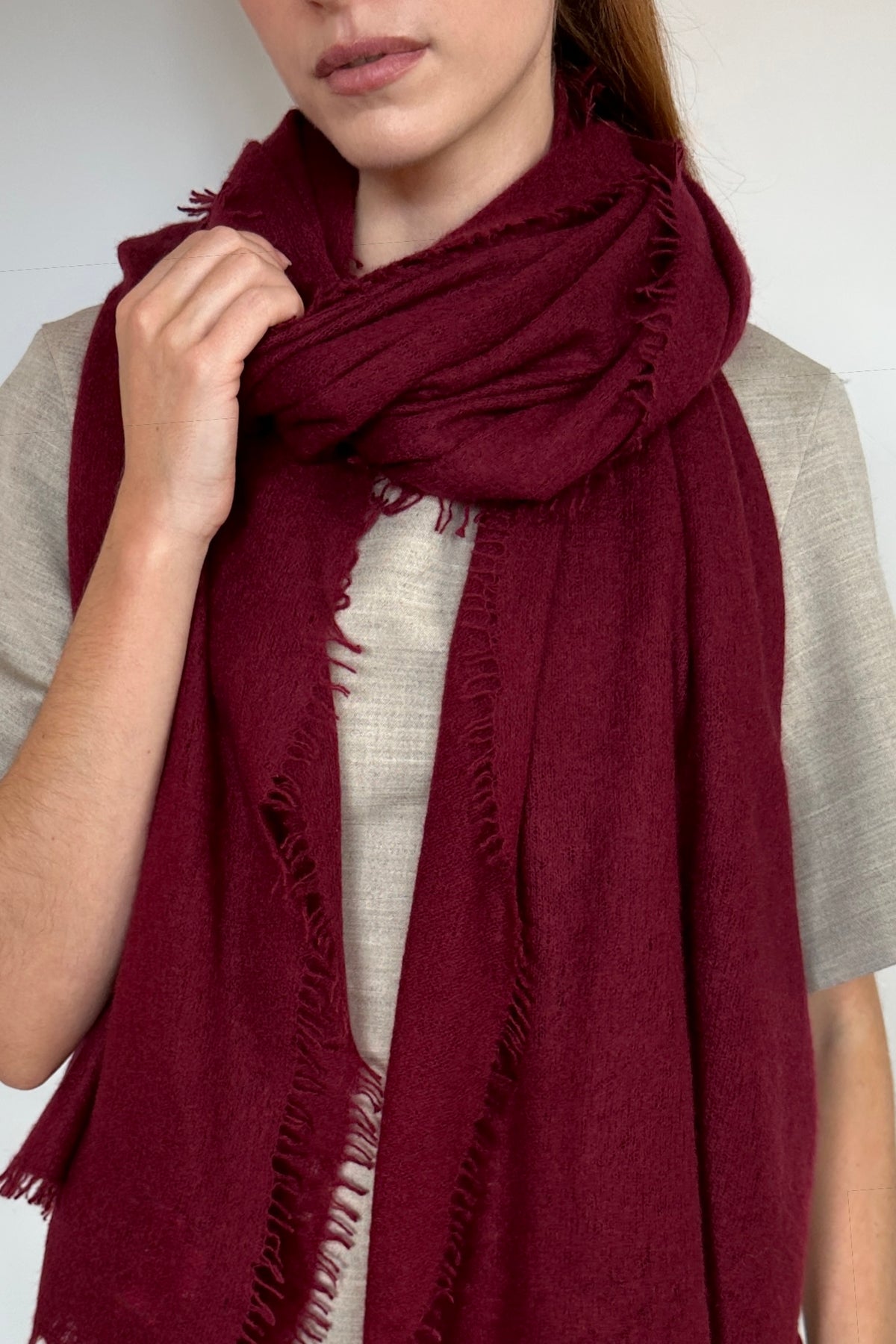 A close-up of a person wearing a deep burgundy cashmere knit scarf with fringed edges over a light beige linen top, against a plain light gray background.