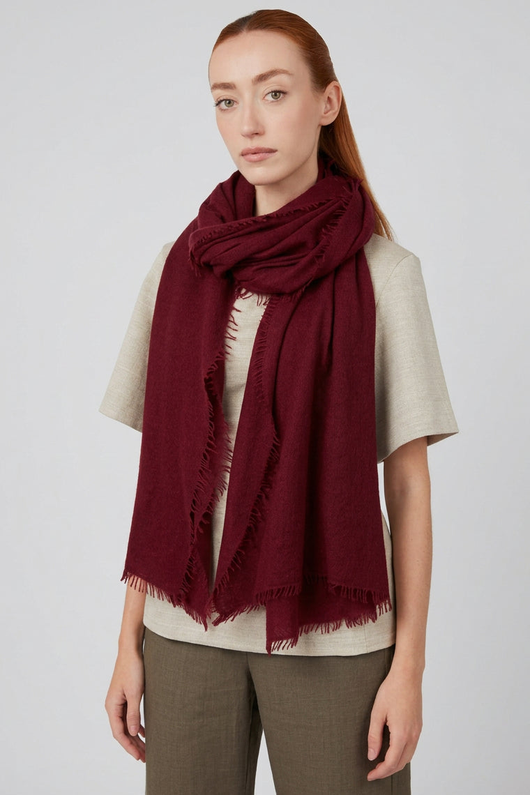 A woman modeling a deep burgundy cashmere knit scarf with fringed edges, styled over a light gray t-shirt and olive green pants against a plain light gray wall.