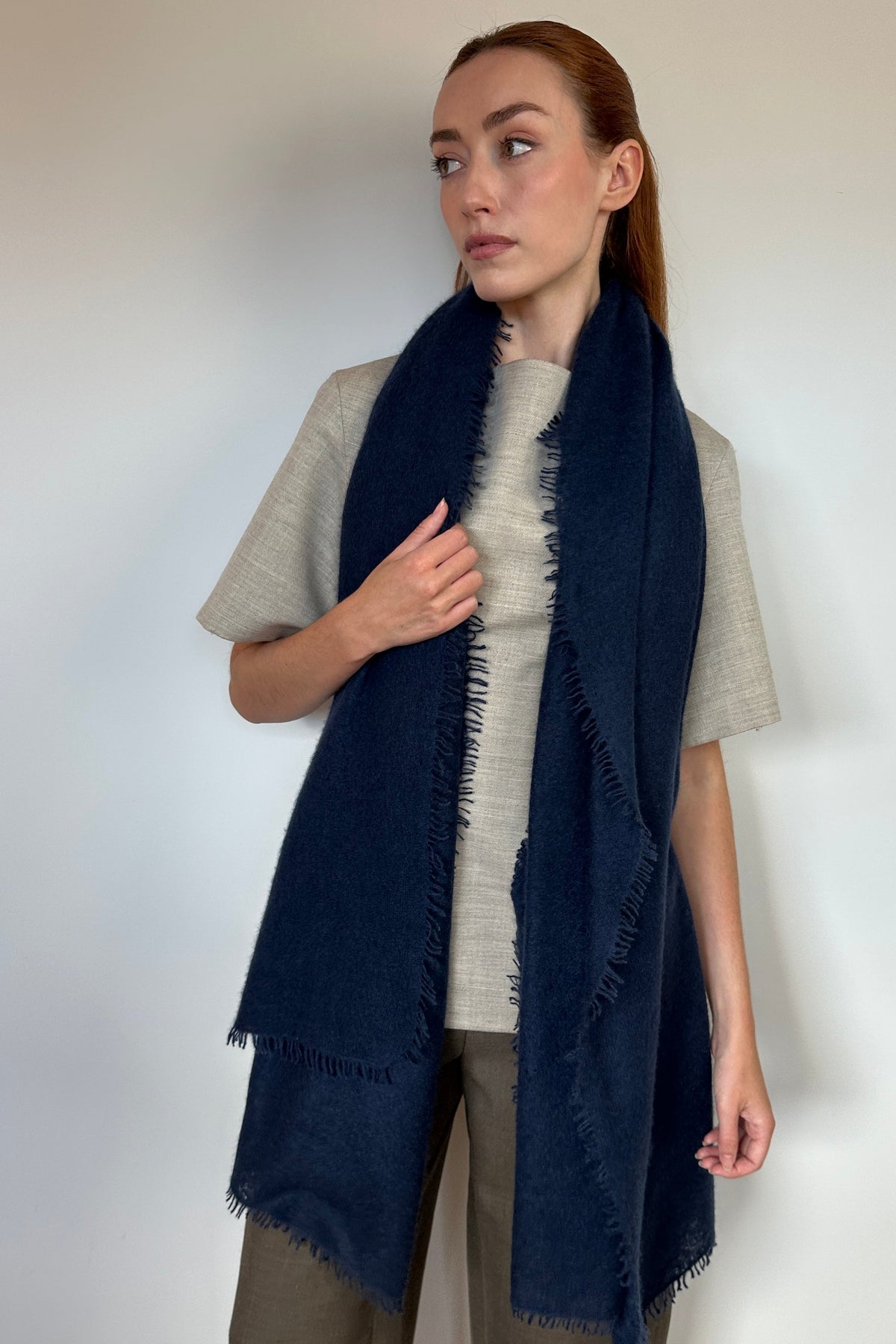 A woman modeling a long navy blue fringed scarf draped over her shoulders, paired with a light gray t-shirt and olive green pants, standing against a plain white wall.