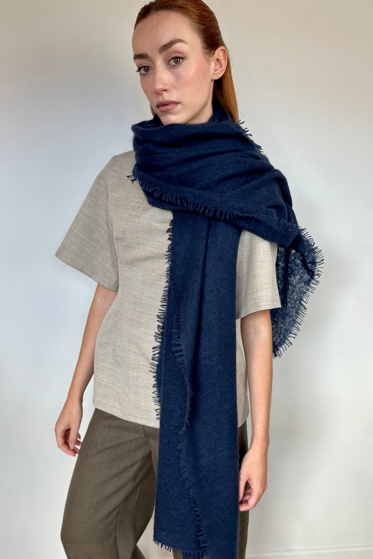 A woman modeling a dark navy blue cashmere fringed scarf wrapped around her neck, paired with a light beige linen top and brown cropped pants against a neutral indoor background.
