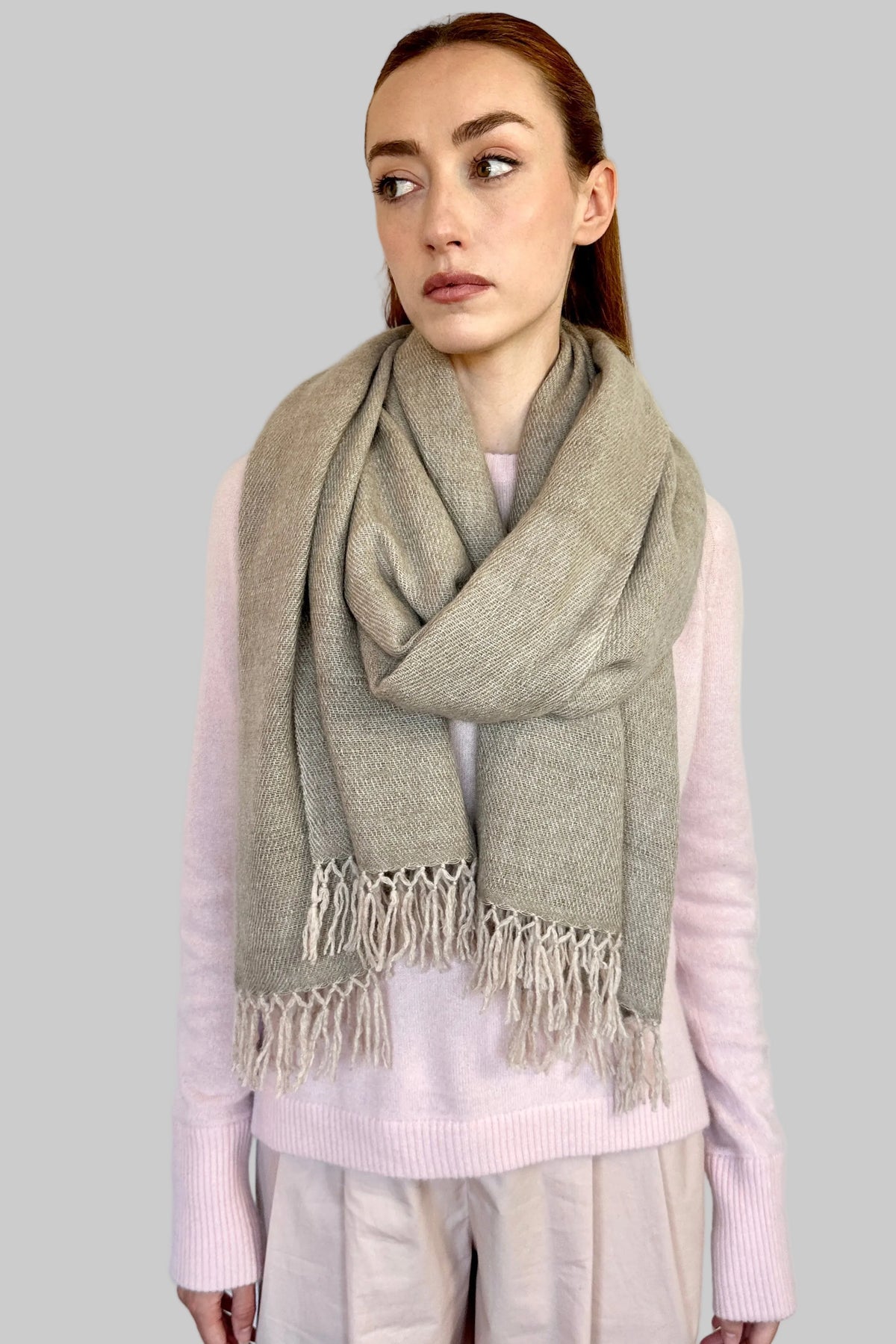 A woman modeling a light beige knit scarf with fringe trim, worn over a pale pink sweater and light beige trousers, standing against a plain off-white wall.