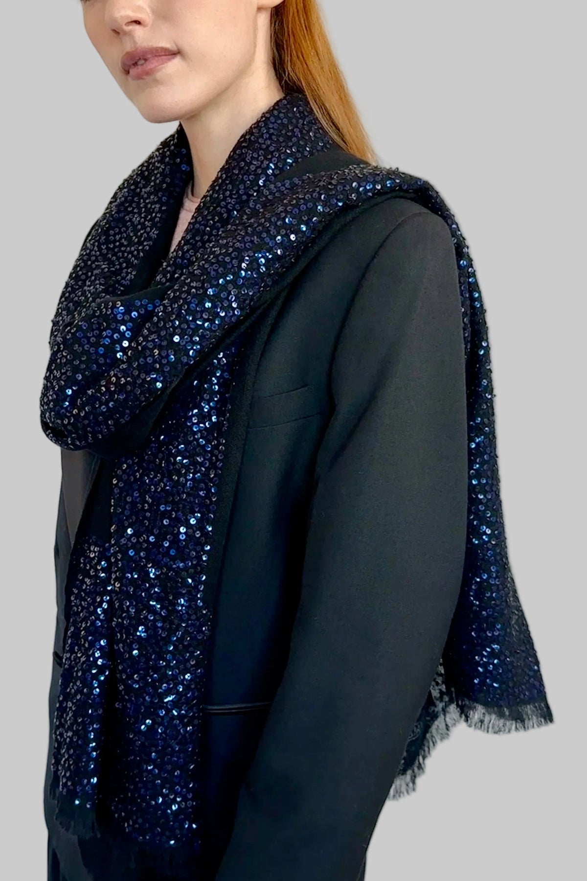 A woman wearing a black blazer and dark pleated skirt, featuring a black base with navy blue sequined cashmere scarf draped over her shoulders, posed against a plain off-white wall.