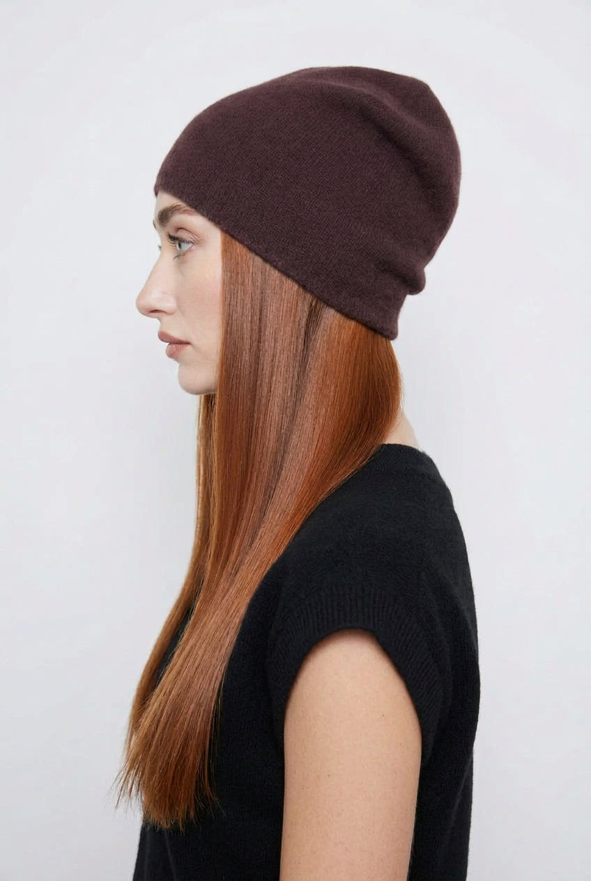 A woman in profile wearing a dark brown slouchy knit beanie and a black short-sleeve top, photographed against a light gray studio background.
