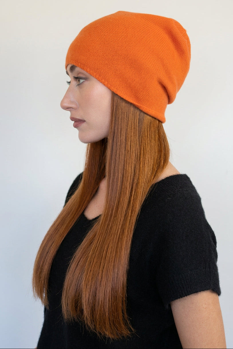 A woman in profile wearing a bright orange knit beanie hat and a black short-sleeve top, photographed against a plain light gray background.