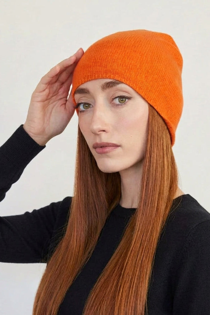 A woman with long auburn hair wearing a bright orange knit cashmere beanie and a black short-sleeve top, posing against a plain off-white wall.