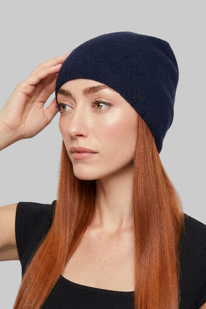 A woman with long red hair wearing a dark navy blue cashmere knit beanie hat and a black top, posing against a light gray background.