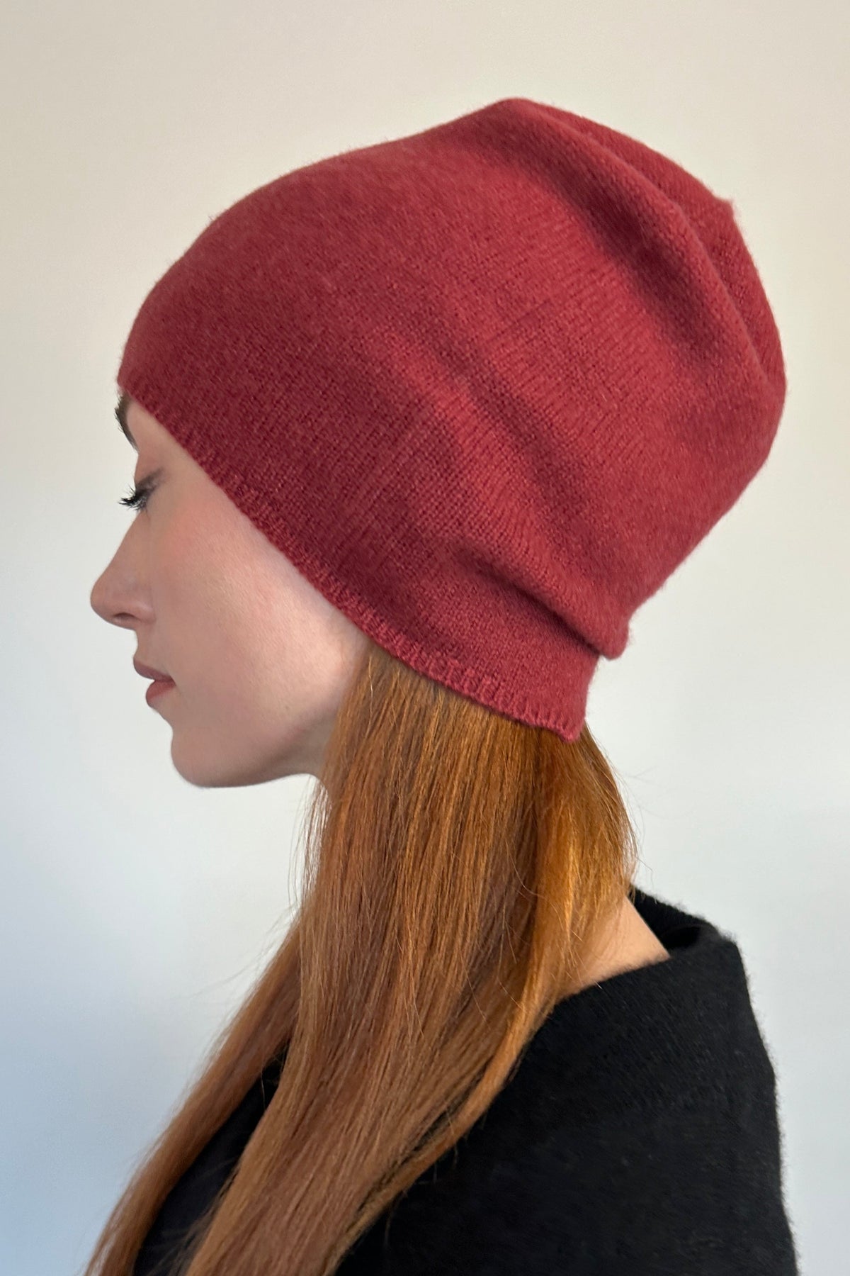A woman shown in profile wearing a ribbed knit beanie hat in a muted brick red color, paired with a black top, against a plain light gray background.