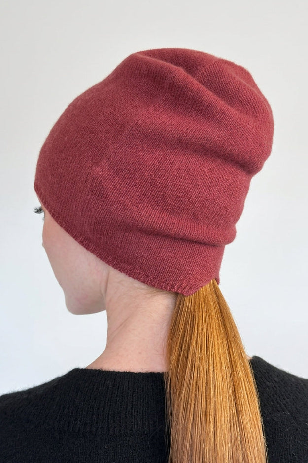 A person shown from the back wearing a slouchy knit beanie in a muted brick red color, with long light brown hair and a black top against a plain light background.