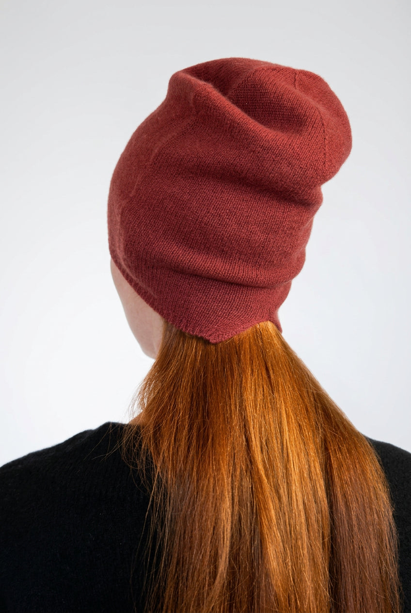 A person shown from the back wearing a slouchy knit beanie in a muted brick red color, with long red hair and a black top against a plain light background.