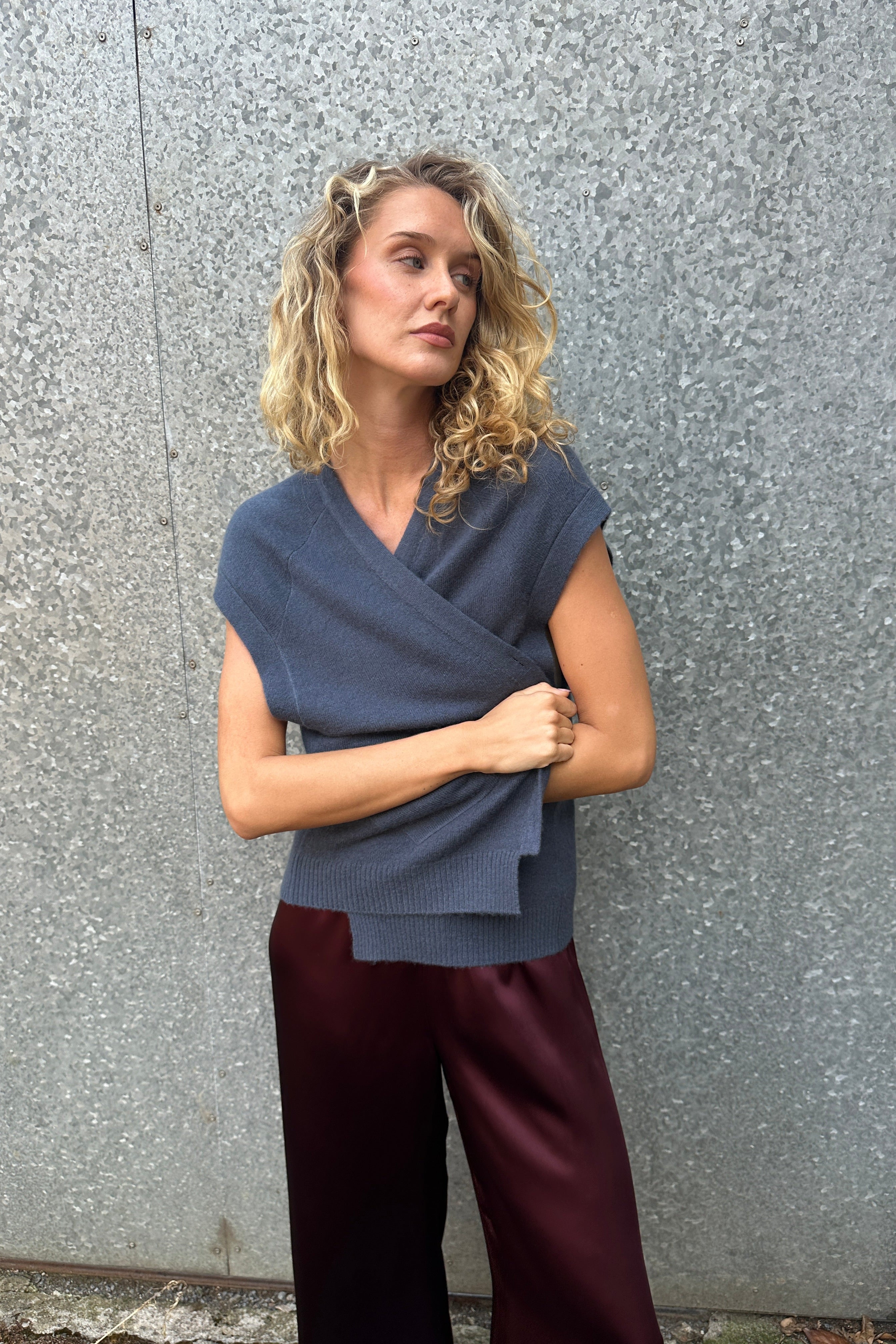 model wearing daisy blue cashmere sleeveless cardigan and plaid scarf against a textured wall.