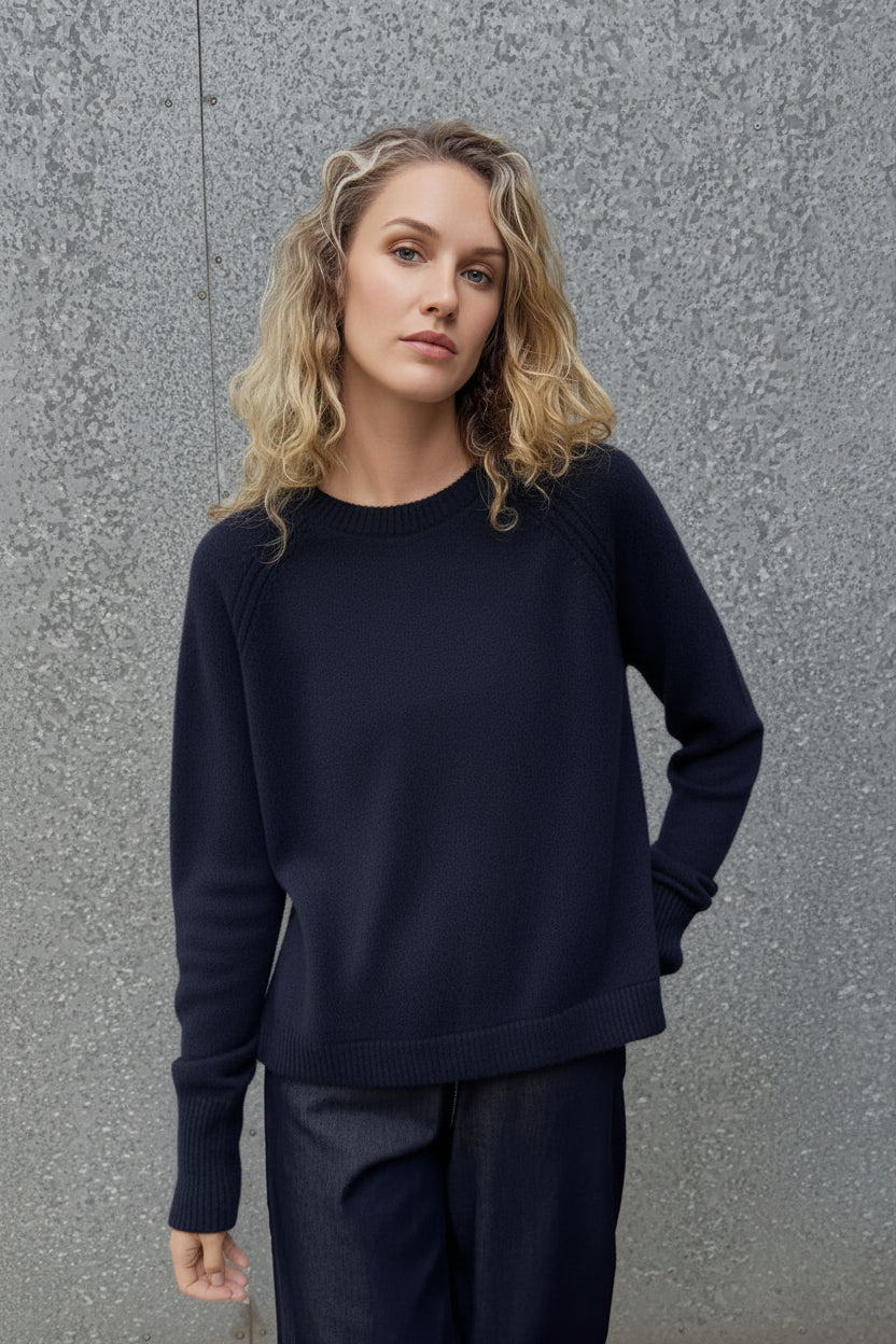 A woman with blonde wavy hair wearing a dark gray crewneck knit sweater and dark blue jeans, standing against a textured gray concrete wall.