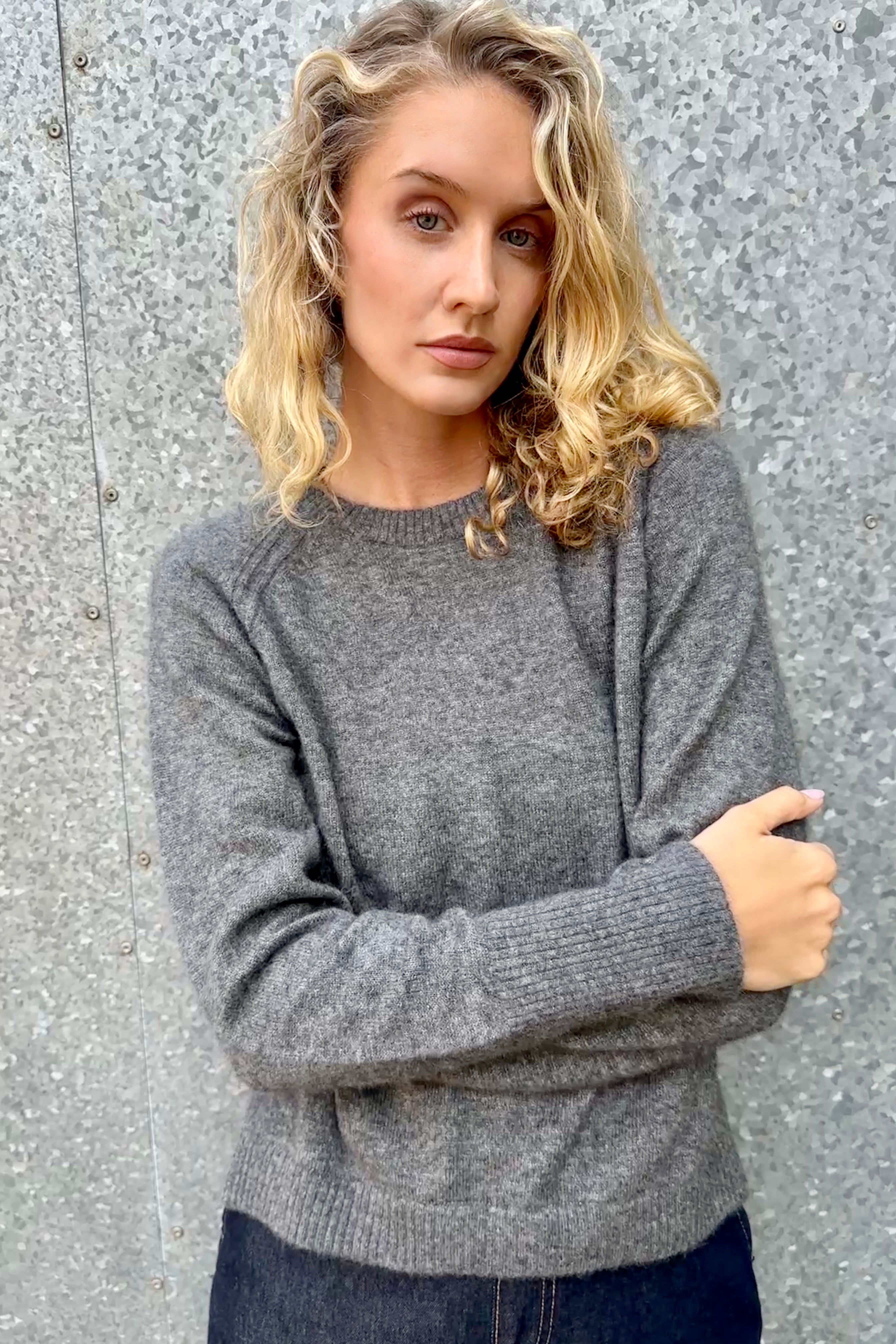 model wearing a gray grey hethna cashmere sweater and dark jeans against a textured wall. close