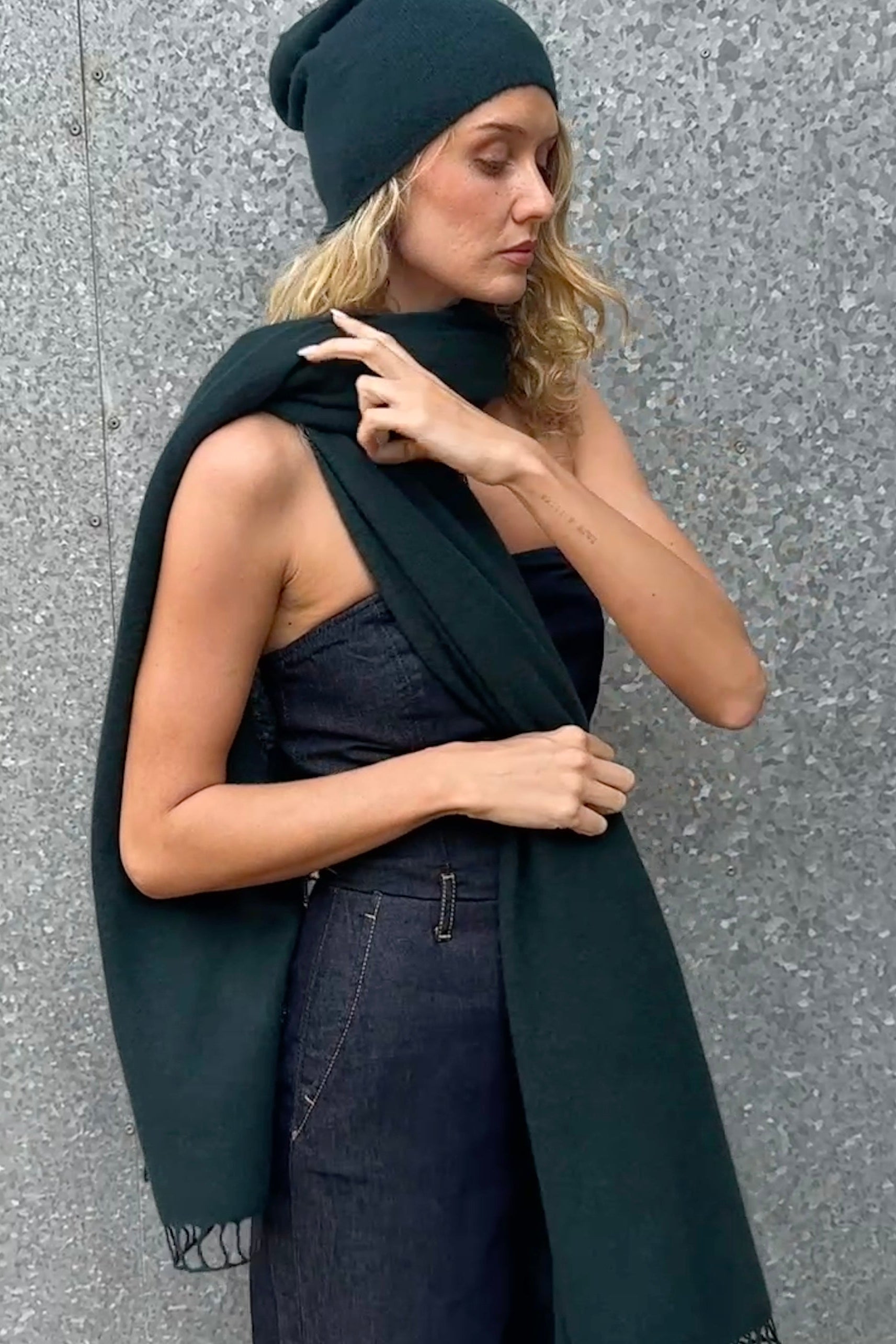 Woman wearing a long dark green scarf and hat over a denim jumpsuit against a gray wall.