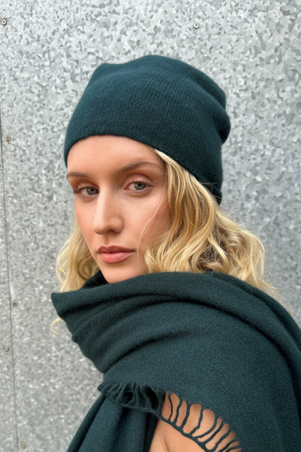 Woman wearing a dark green beanie and scarf against a textured wall.