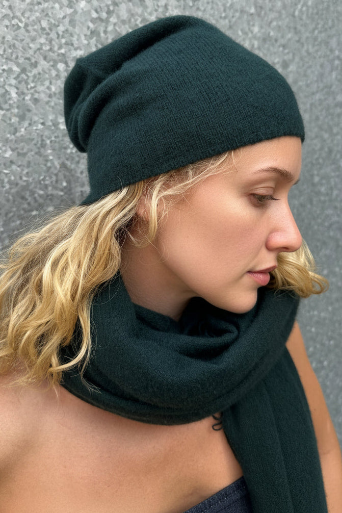 Woman wearing a dark green beanie and scarf against a textured gray wall