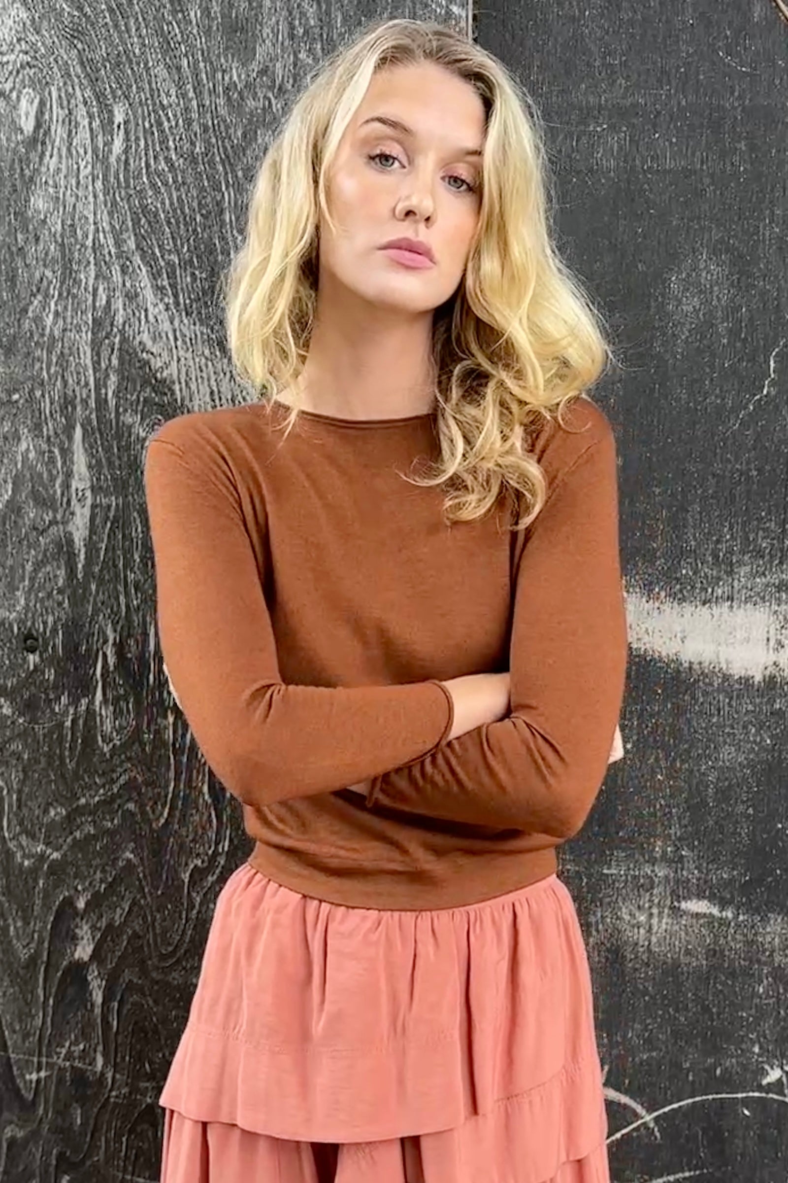 model wearing a rust brown cashmere silk top or sweater and pink skirt against a textured black wall