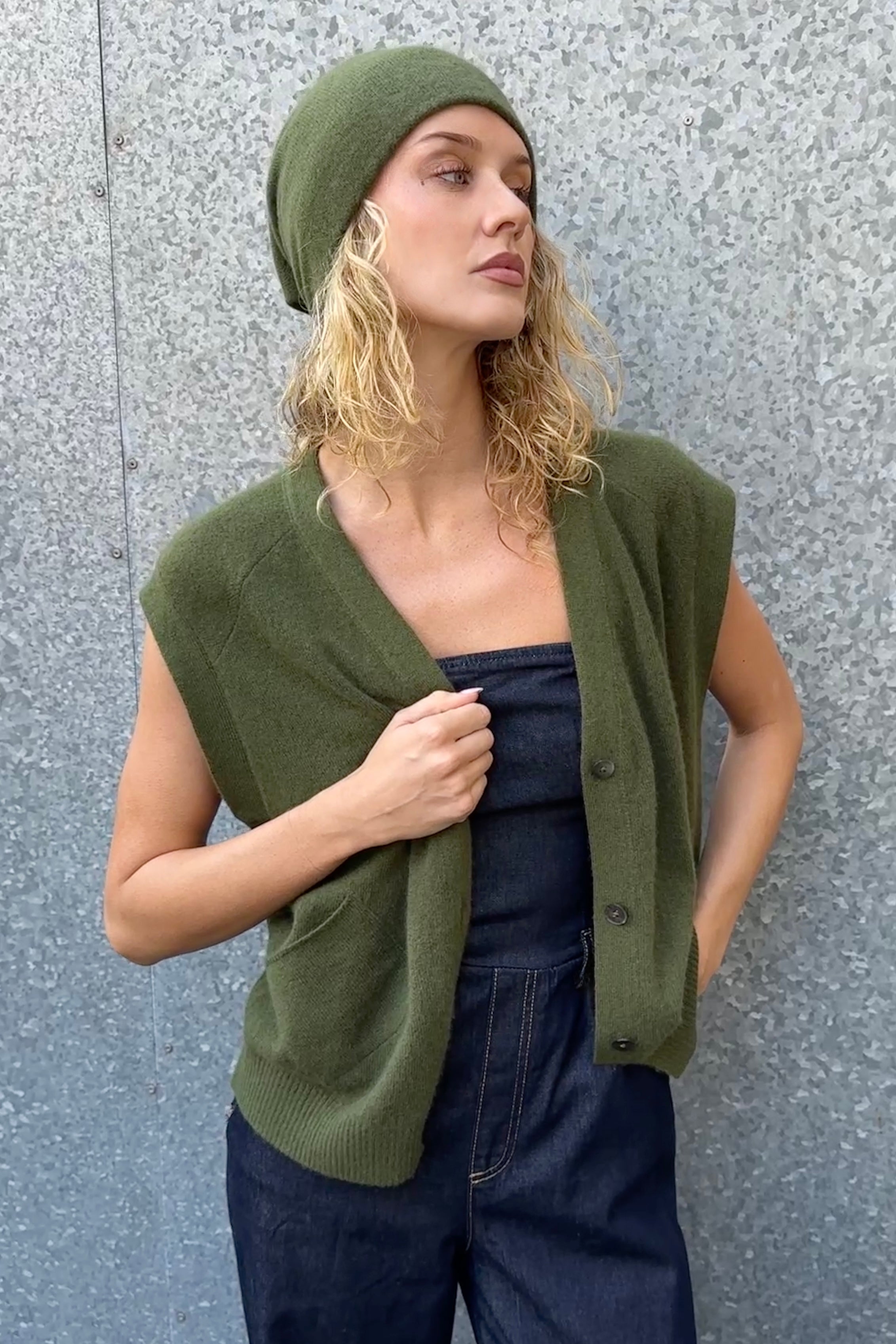 Woman wearing a green knitted vest and beanie against a gray wall
