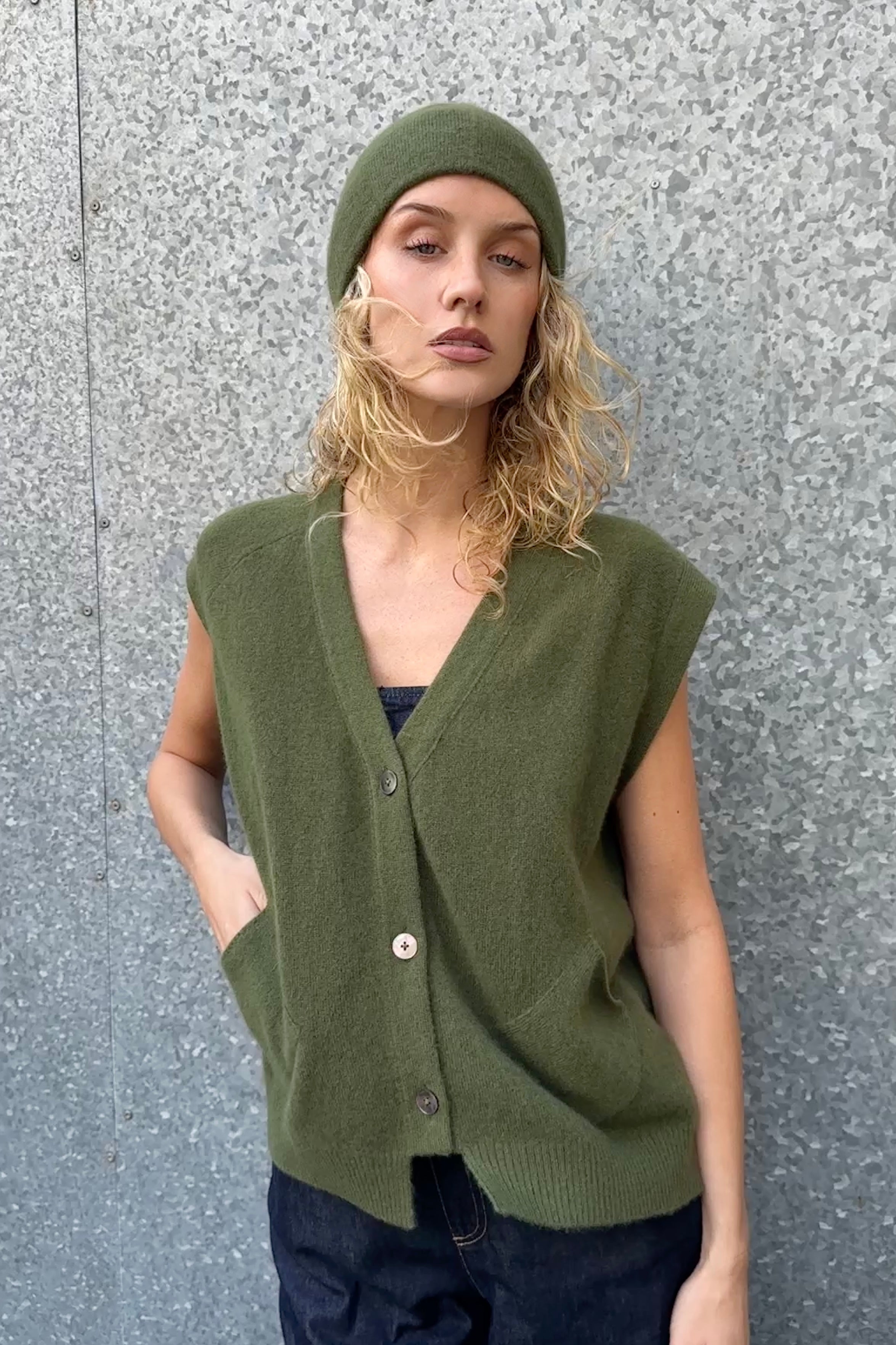 Woman wearing a green knitted vest and beanie against a gray wall