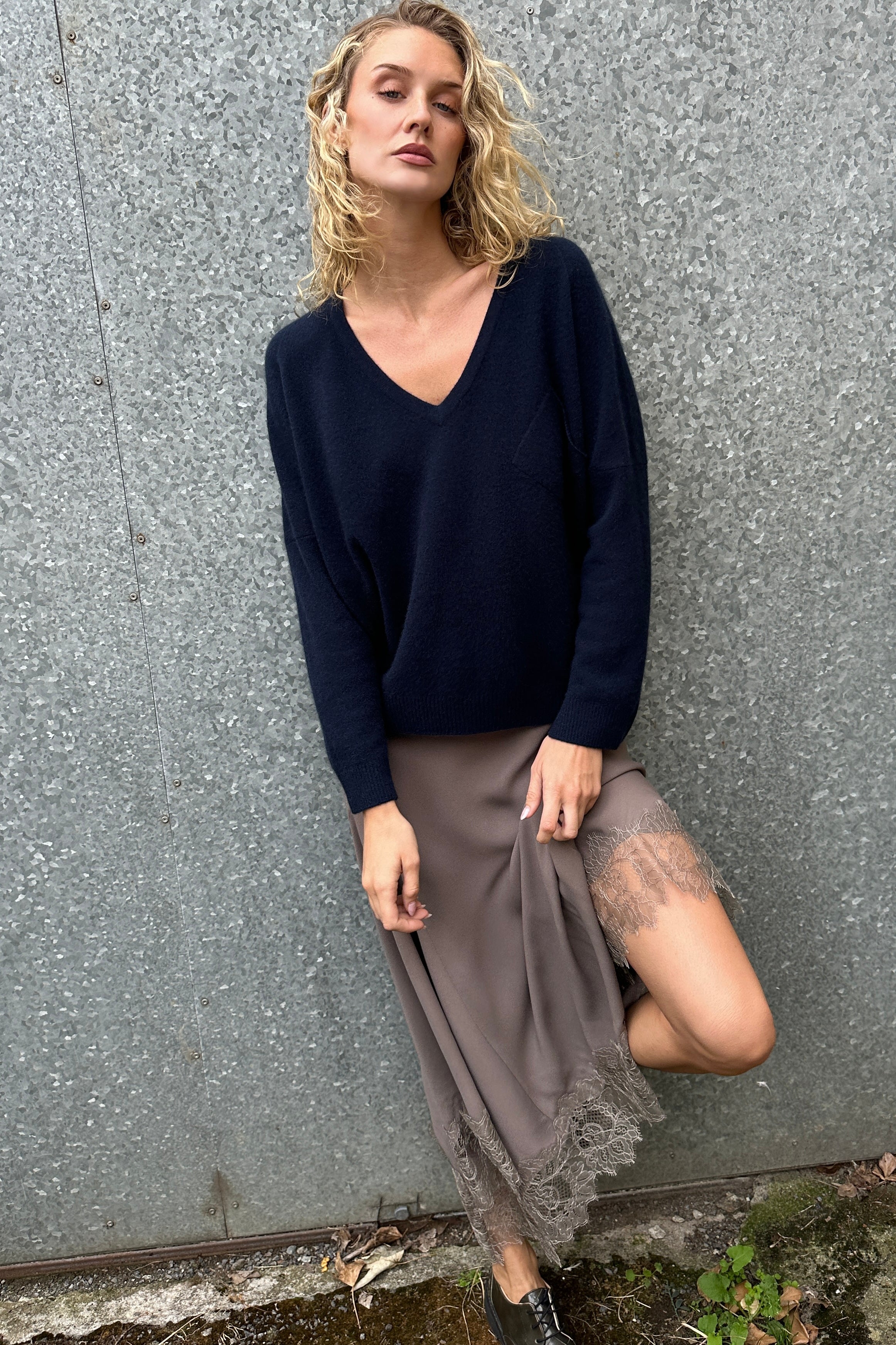 Woman wearing a navy blue v neck sweater and gray skirt against a textured wall.