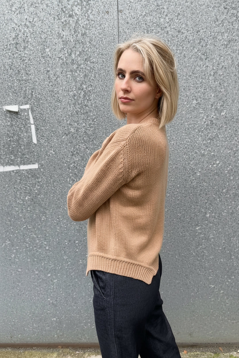 Woman wearing a beige sweater and dark pants against a gray wall.