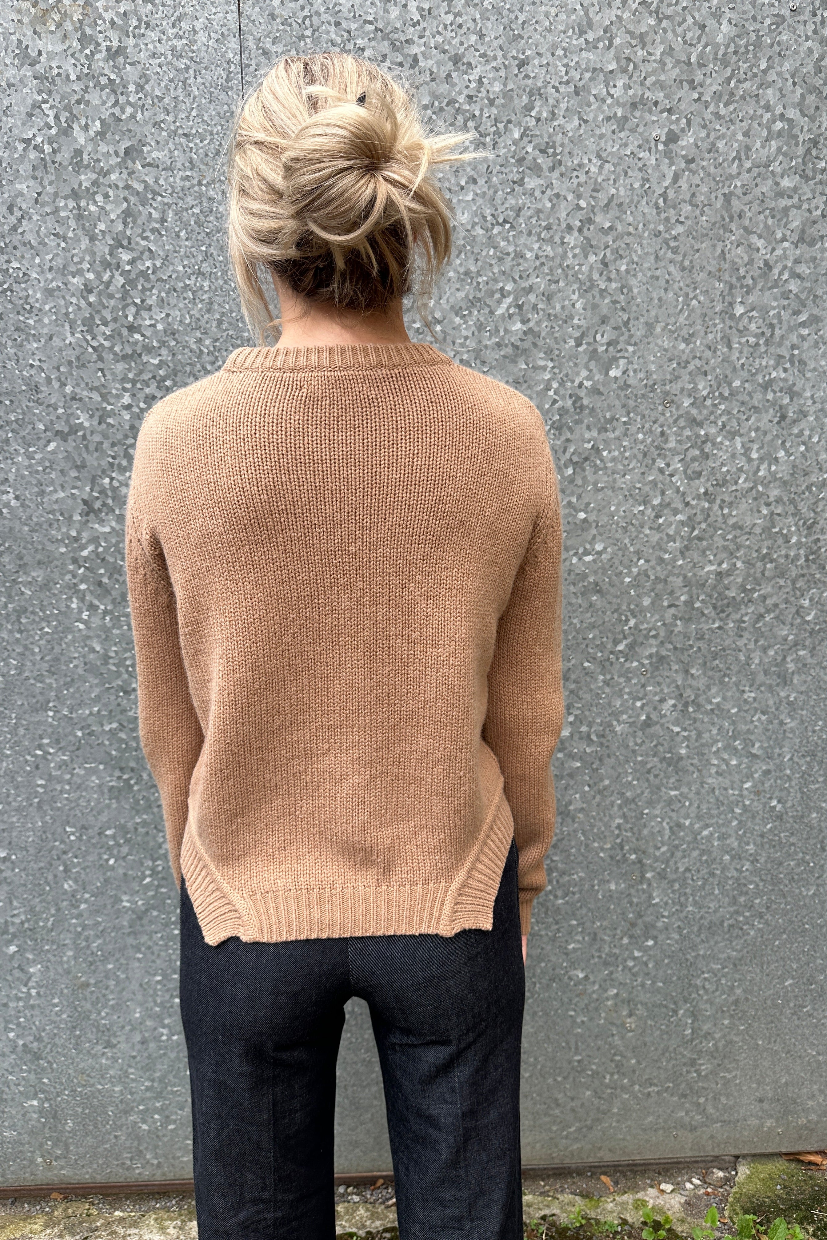 Woman wearing a beige sweater standing against a gray wall. bakc view
