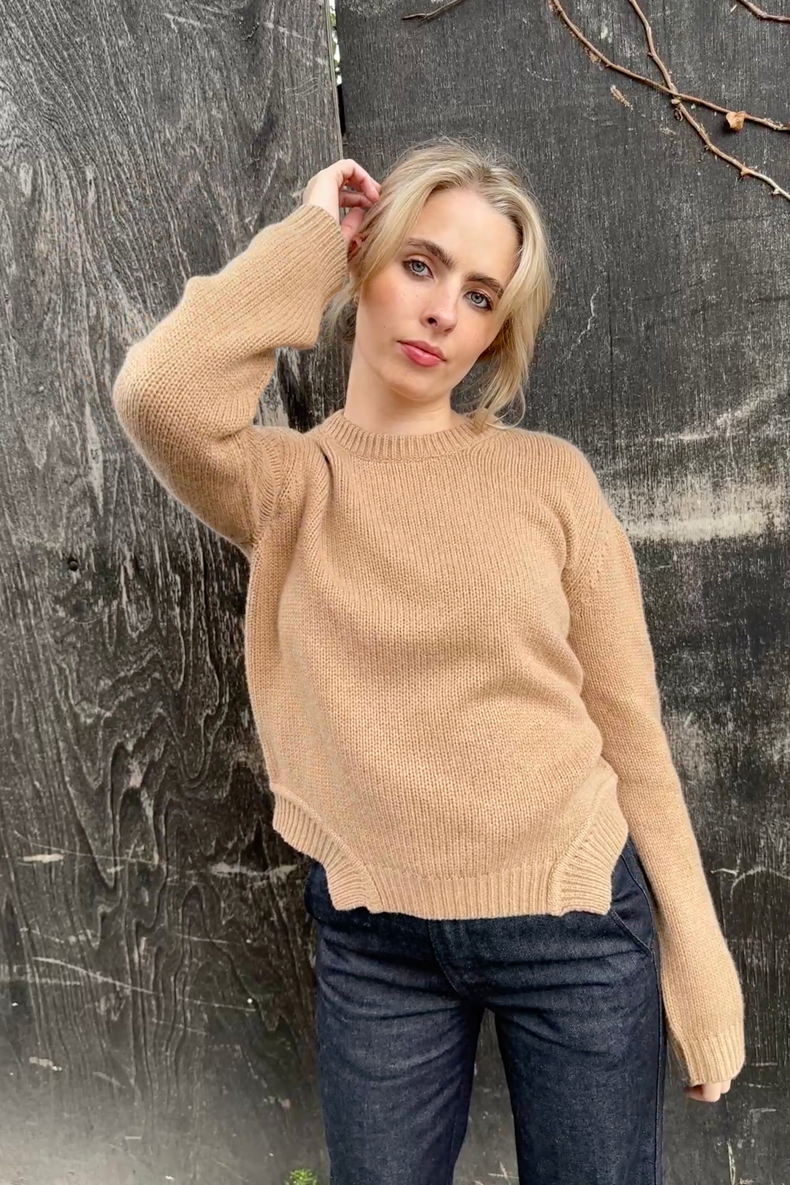 Woman wearing a beige sweater and dark jeans against a textured wall.