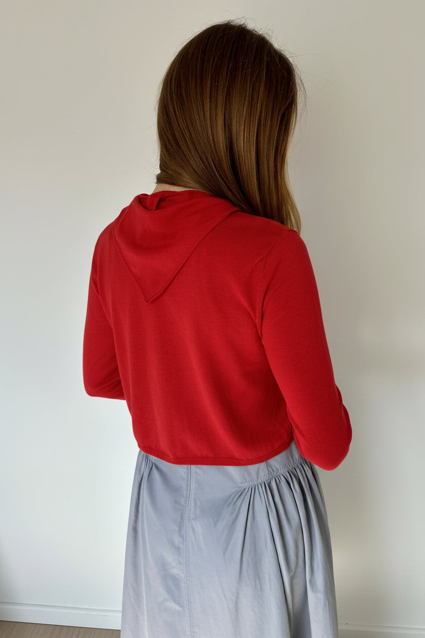 A woman viewed from behind wearing a red cardi and a dusty blue skirt, standing against a plain off-white wall.