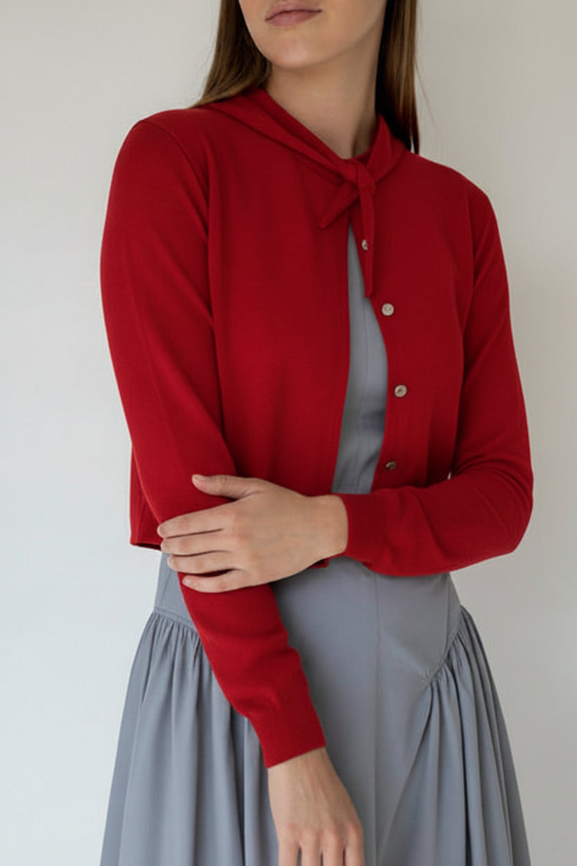 A woman wearing a
red cropped cardigan with a tie-neck detail over a dusty blue dress, standing against a plain off-white wall.
