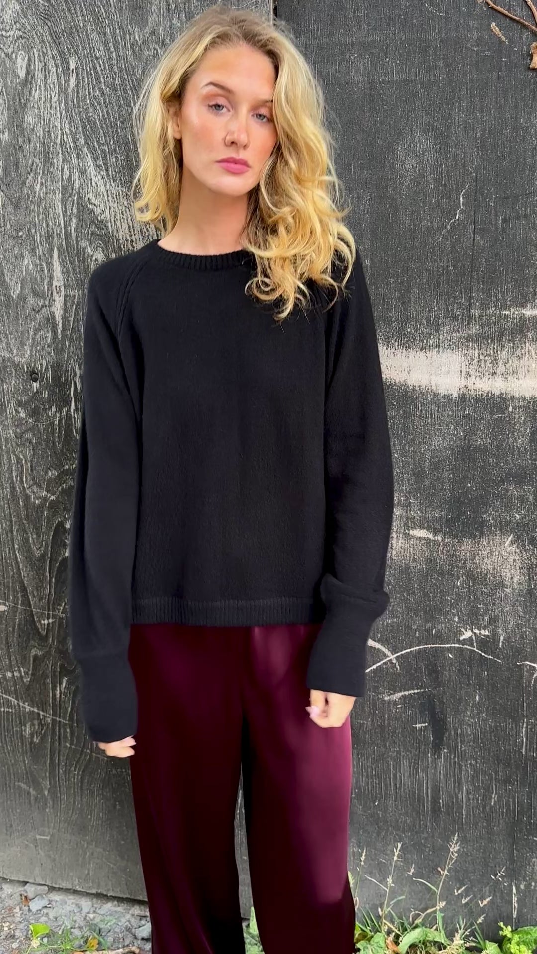 Woman wearing a black cashmere sweater and burgundy pants standing against a textured wall.